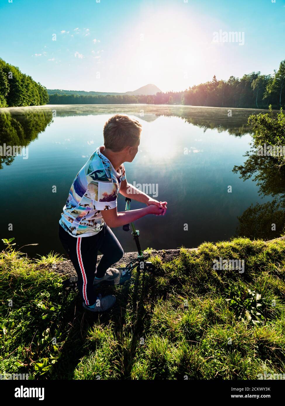Un garçon de cheveux blond avec un scooter se tient sur un sentier de parc le long du lac. Concept de sports d'enfance. Le soleil du soir fait des éruptions et des réflexions dans l'eau. Banque D'Images