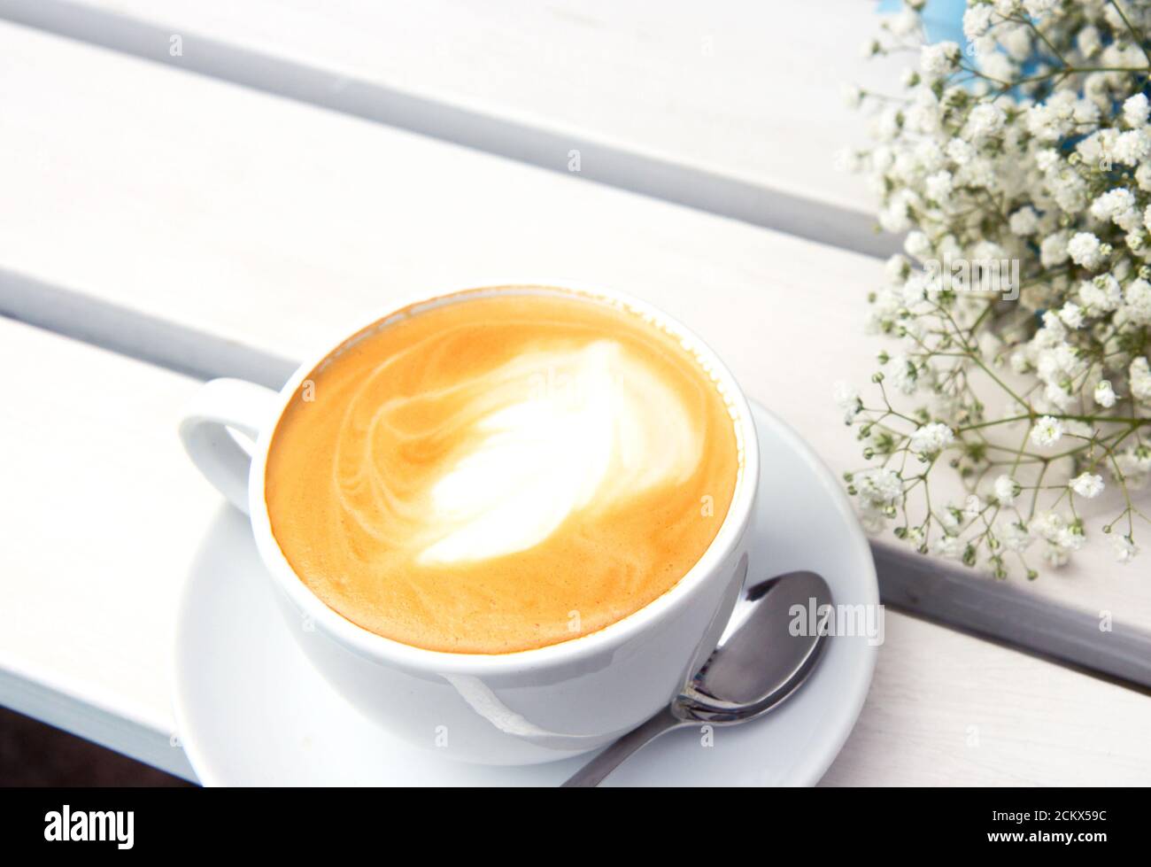 Cappuccino dans une tasse en céramique blanche et bouquet de petites fleurs blanches sur un banc en bois. Café rustique en plein air le matin. Routine du matin. Gypsophila Banque D'Images