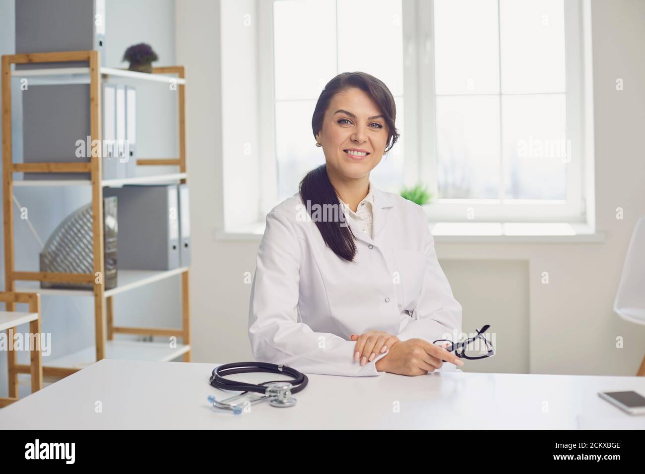 Femme médecin pratiquant regardant la caméra tout en étant assis à une table dans un bureau de clinique. Banque D'Images