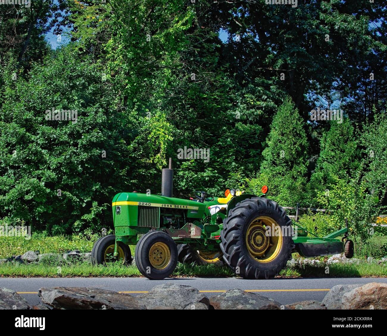 Un tracteur John Deere classique avec une plate-forme de tonte fixée à l'arrière est stationné sur le côté de la route. En arrière-plan se trouvent des arbres verts luxuriants Banque D'Images