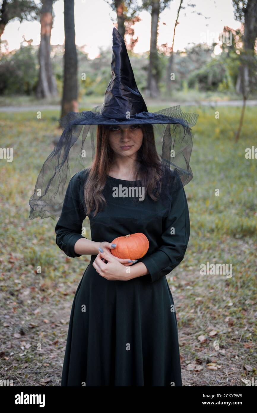 Une jeune femme en robe noire et chapeau de sorcière tient une citrouille dans ses mains. Costume de fête d'Halloween. Forêt, parc avec arbres d'automne Banque D'Images