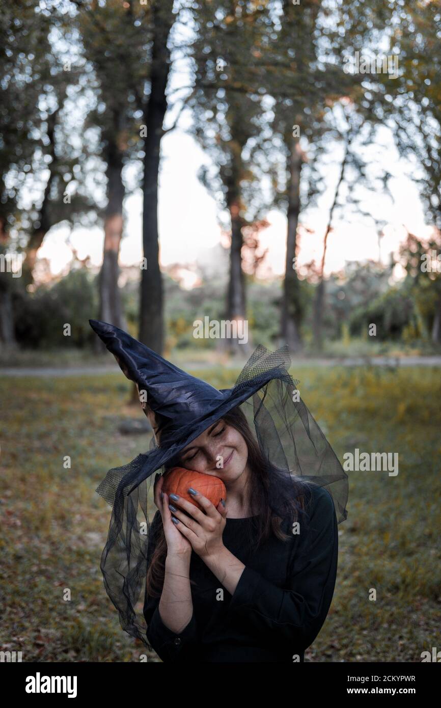 Une jeune femme en robe noire et chapeau de sorcière tient une citrouille dans ses mains. Costume de fête d'Halloween. Forêt, parc avec arbres d'automne Banque D'Images