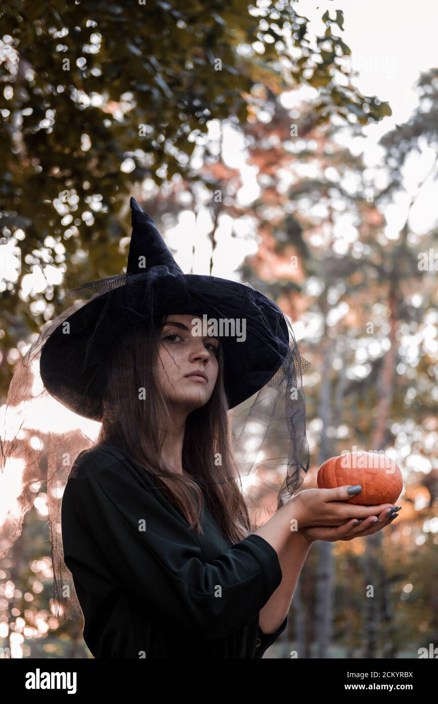 Une jeune femme en robe noire et chapeau de sorcière tient une citrouille dans ses mains. Costume de fête d'Halloween. Forêt, parc avec arbres d'automne Banque D'Images