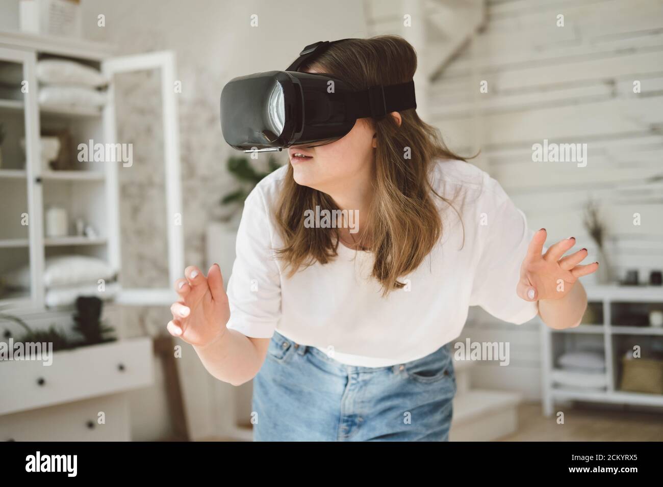 Jeune femme en jeans et T-shirt blanc portant le casque de réalité virtuelle joue au jeu, travaille, regarde la vidéo dans la chambre à côté du lit. Concept de technolo moderne Banque D'Images