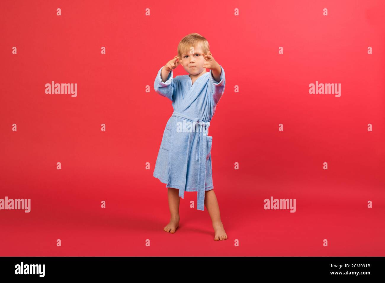 Un enfant blond garçon en peignoir bleu sur un fond rouge vif montre un signe de la main tire un pistolet ou cible de buts près des yeux. Photo studio d'un enfant dansant Banque D'Images