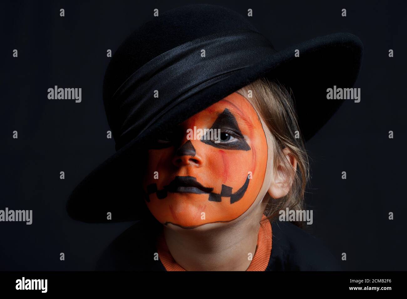 Triste enfant avec un motif de citrouille sur son visage dans le chapeau noir sur un fond noir, Halloween et ressemble à un Joker Banque D'Images