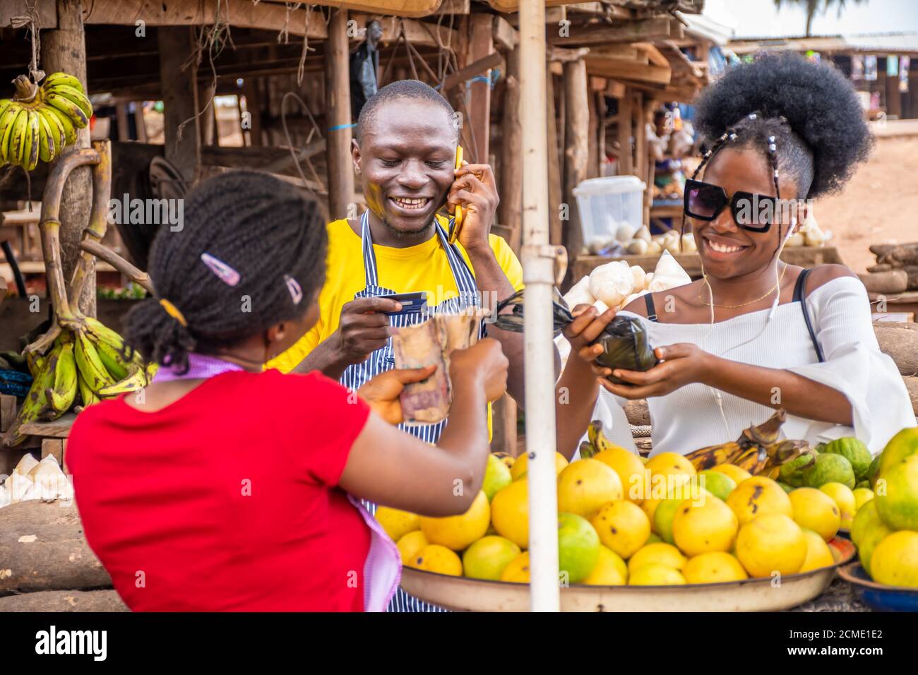 jeune homme noir faisant un appel téléphonique et tenant une carte de crédit, femme payant pour de la nourriture dans un marché africain local Banque D'Images