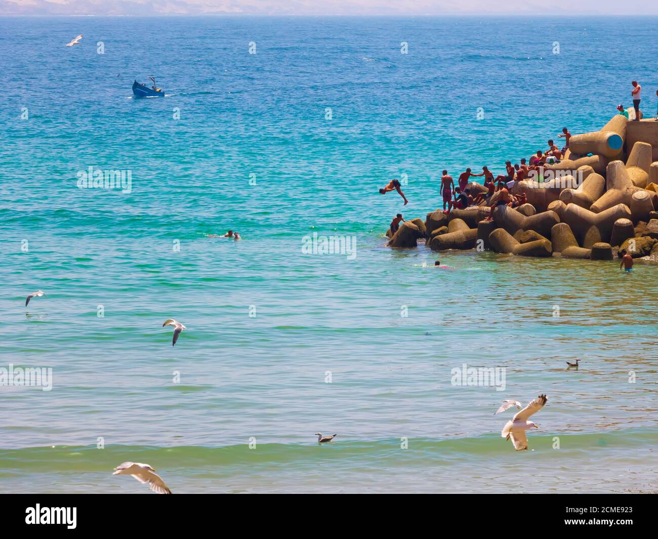 Agadir, Maroc - 14 AOÛT 2020 : personnes nageant au bord d'un port, dans l'océan Atlantique bleu, avec des mouettes au-dessus et un bateau bleu Banque D'Images Agadir, Maroc - 14 AOÛT 2020 : personnes nageant au bord d'un port, dans l'océan Atlantique bleu, avec des mouettes au-dessus et un bateau bleu Banque D'Images