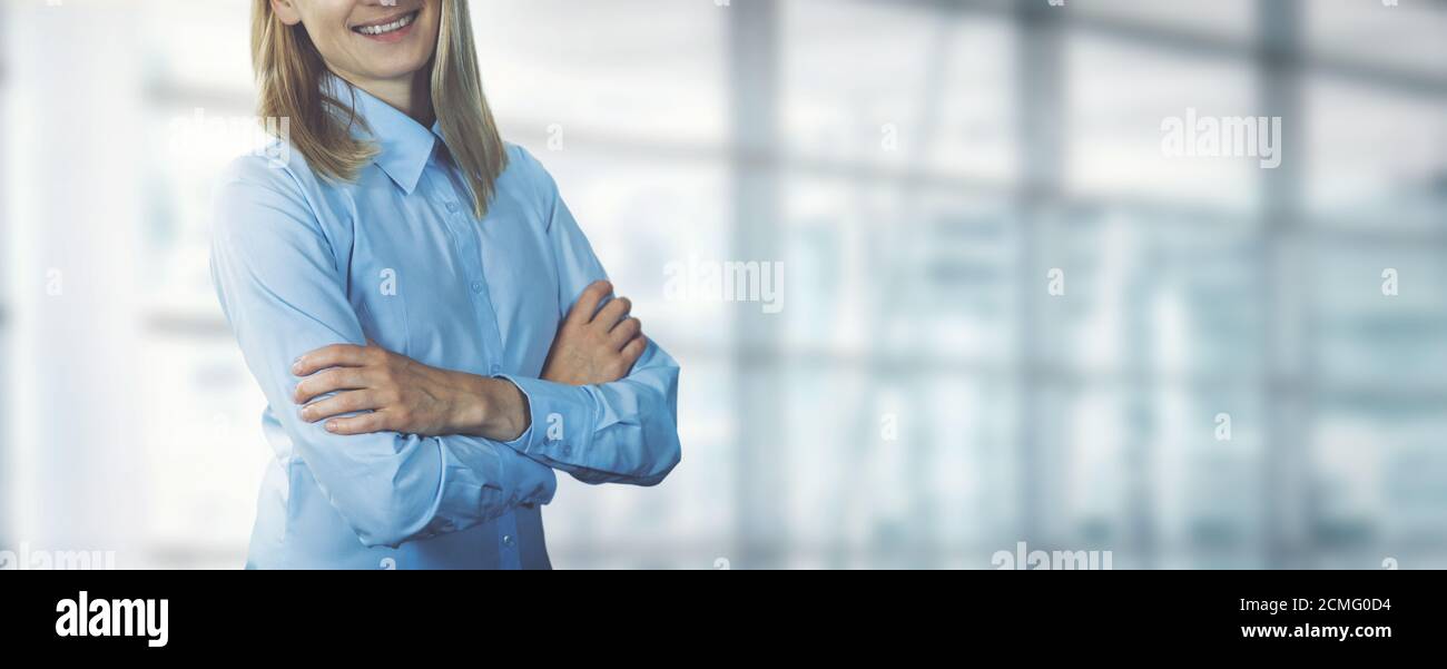 jeune femme d'affaires souriante portant un chemisier bleu debout avec les bras croisés dans un bureau moderne. espace de copie de bannière Banque D'Images