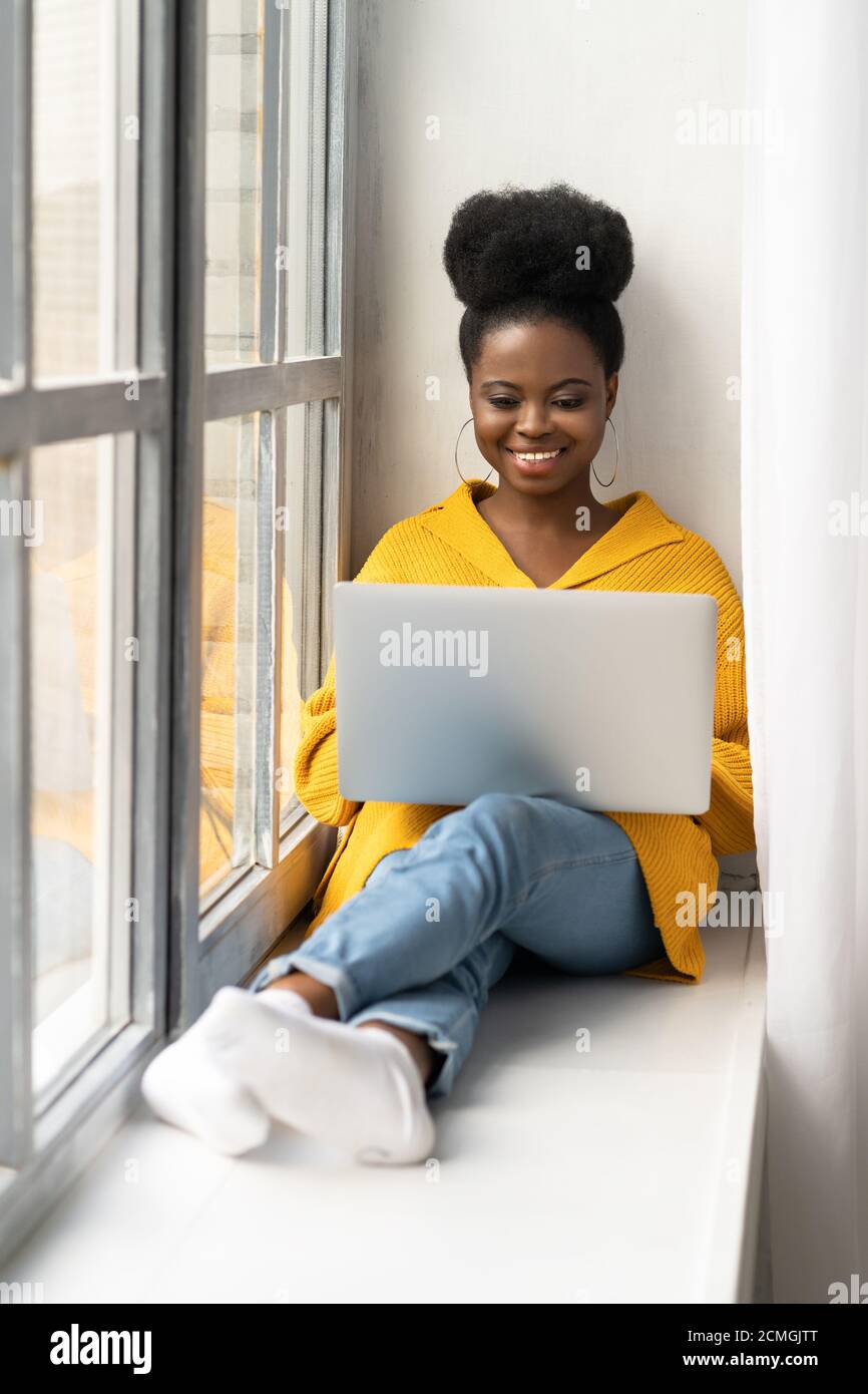 Jeune femme biracial afro-américaine souriante avec une coiffure afro-américaine dans un gilet jaune assis sur un rebord de fenêtre, travaillant à distance sur un ordinateur portable, bavardant et t Banque D'Images