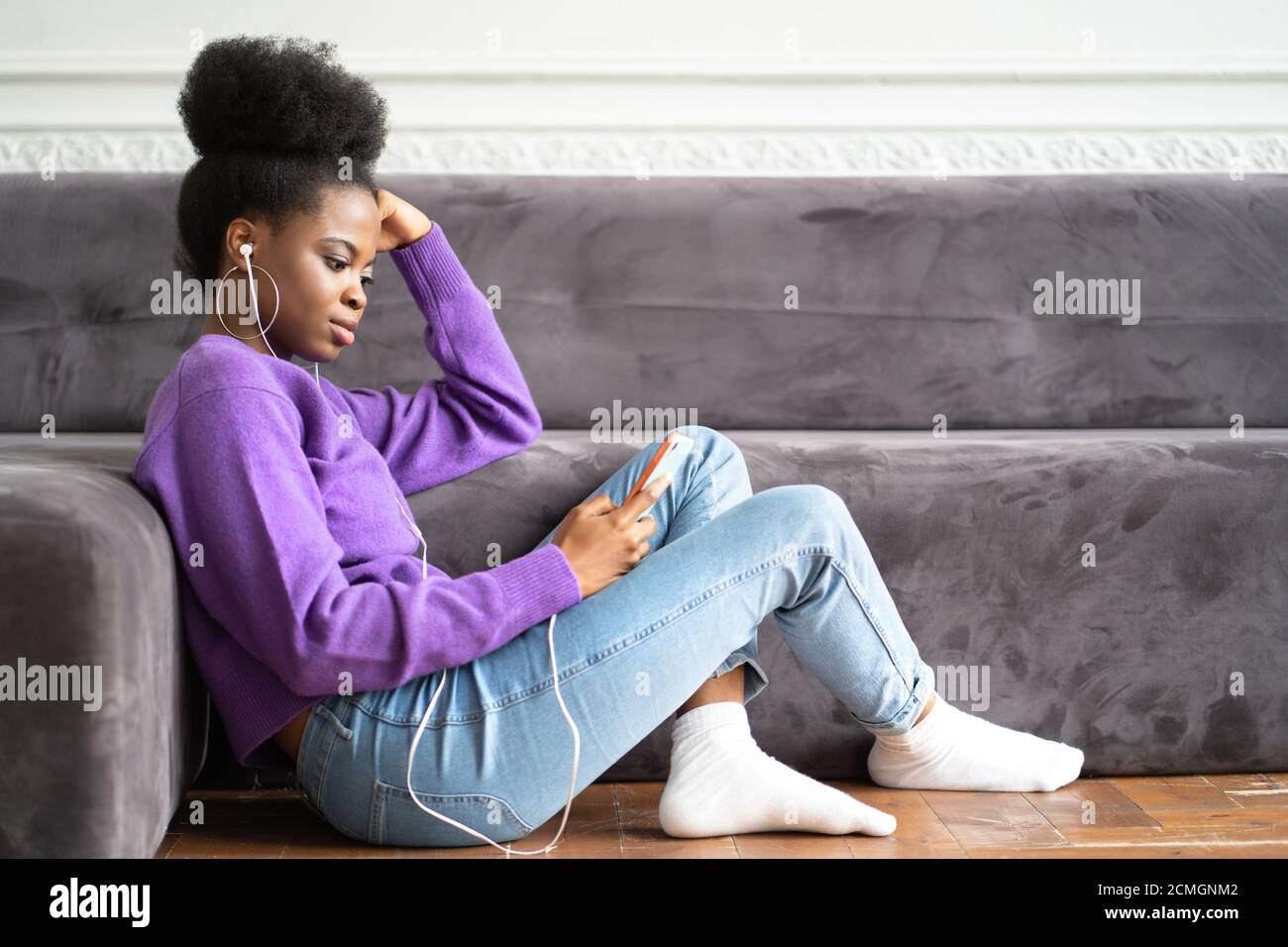 Jeune femme millénaire afro-américaine dans un chandail violet assis sur le sol, en utilisant un smartphone, en tapant un message, écoute de la musique, défile à travers Banque D'Images