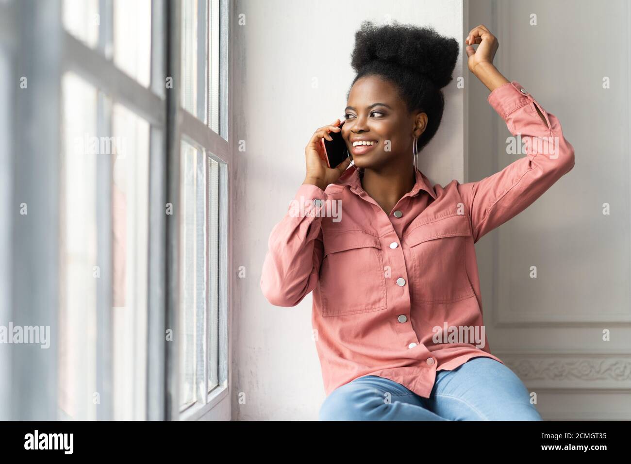 Bonne femme africaine américaine millénaire avec afro coiffure porter chemise rose, assis sur le rebord de la fenêtre, souriant, prenant le téléphone, regardant la fenêtre. Noir Banque D'Images