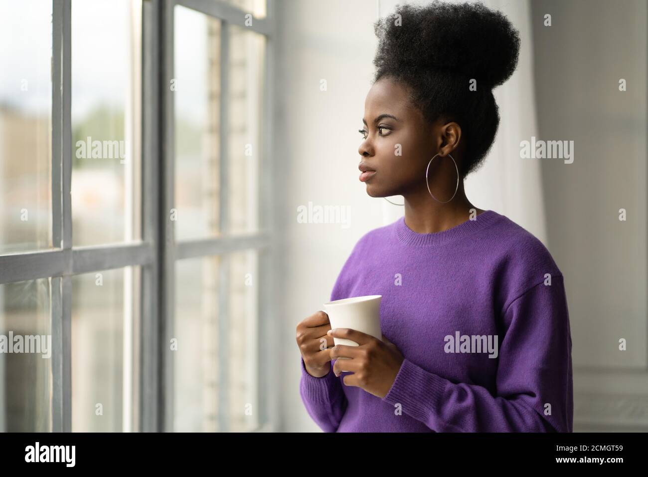 Pensive Afro-américaine jeune femme millénaire dans les bijoux à la mode grandes boucles d'oreilles porter le chandail violet pensée, regardant à travers la fenêtre, tenant blanc Banque D'Images