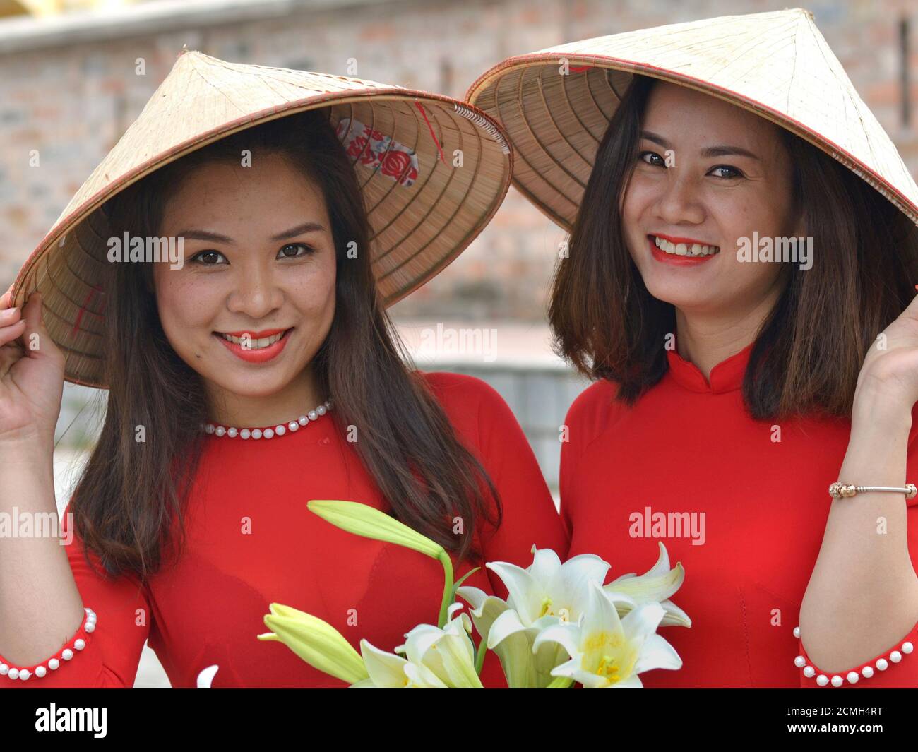 Deux jeunes femmes vietnamiennes portent des chapeaux coniques asiatiques  et des robes vietnamiennes rouges, tiennent son bord de leurs chapeaux avec  leurs mains, et souriez pour l'appareil photo Photo Stock - Alamy