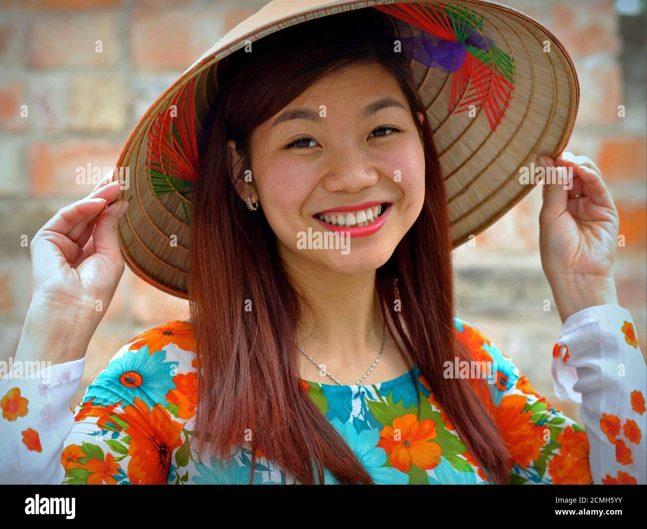 Belle jeune femme vietnamienne avec de longs cheveux tient le bord de son chapeau conique asiatique avec les deux mains et sourires à l'appareil photo. Banque D'Images