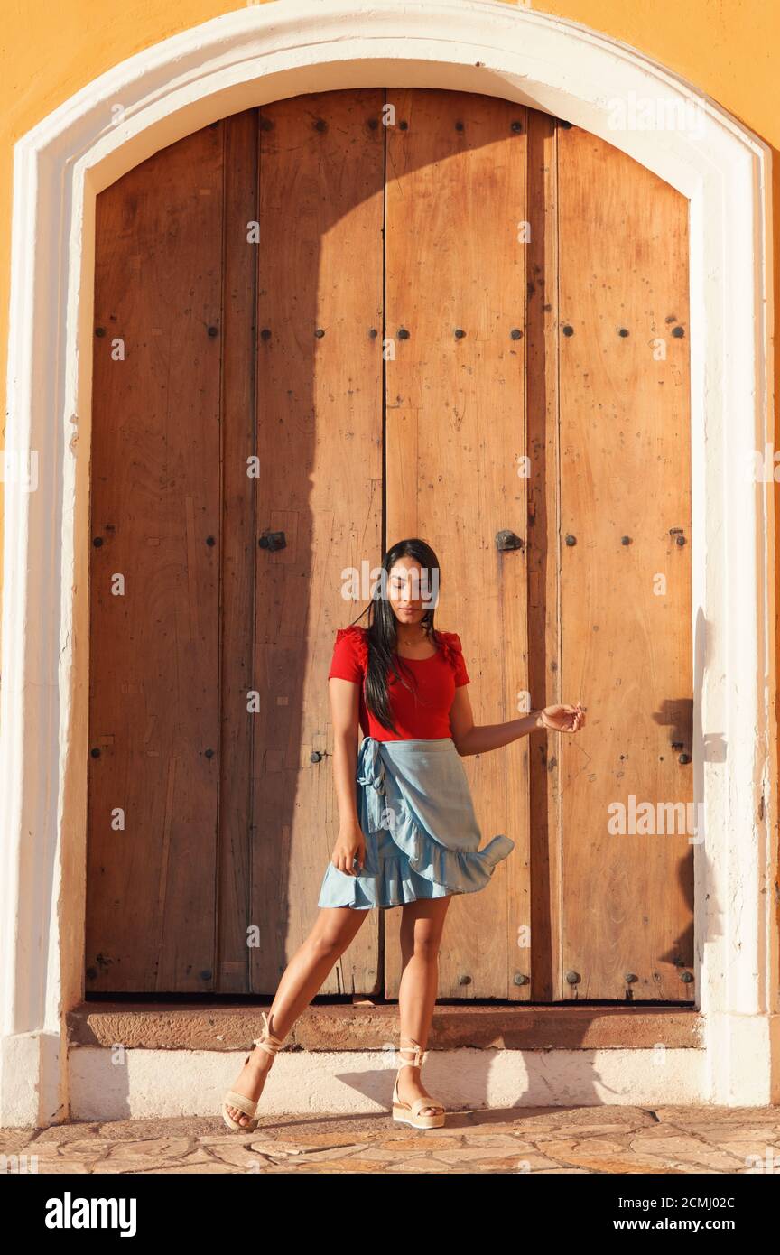 Une jeune femme en été portant une chemise rouge, une jupe bleu vif et des sandales à espadrilles beiges qui se posent à côté d'une grande porte vintage Banque D'Images