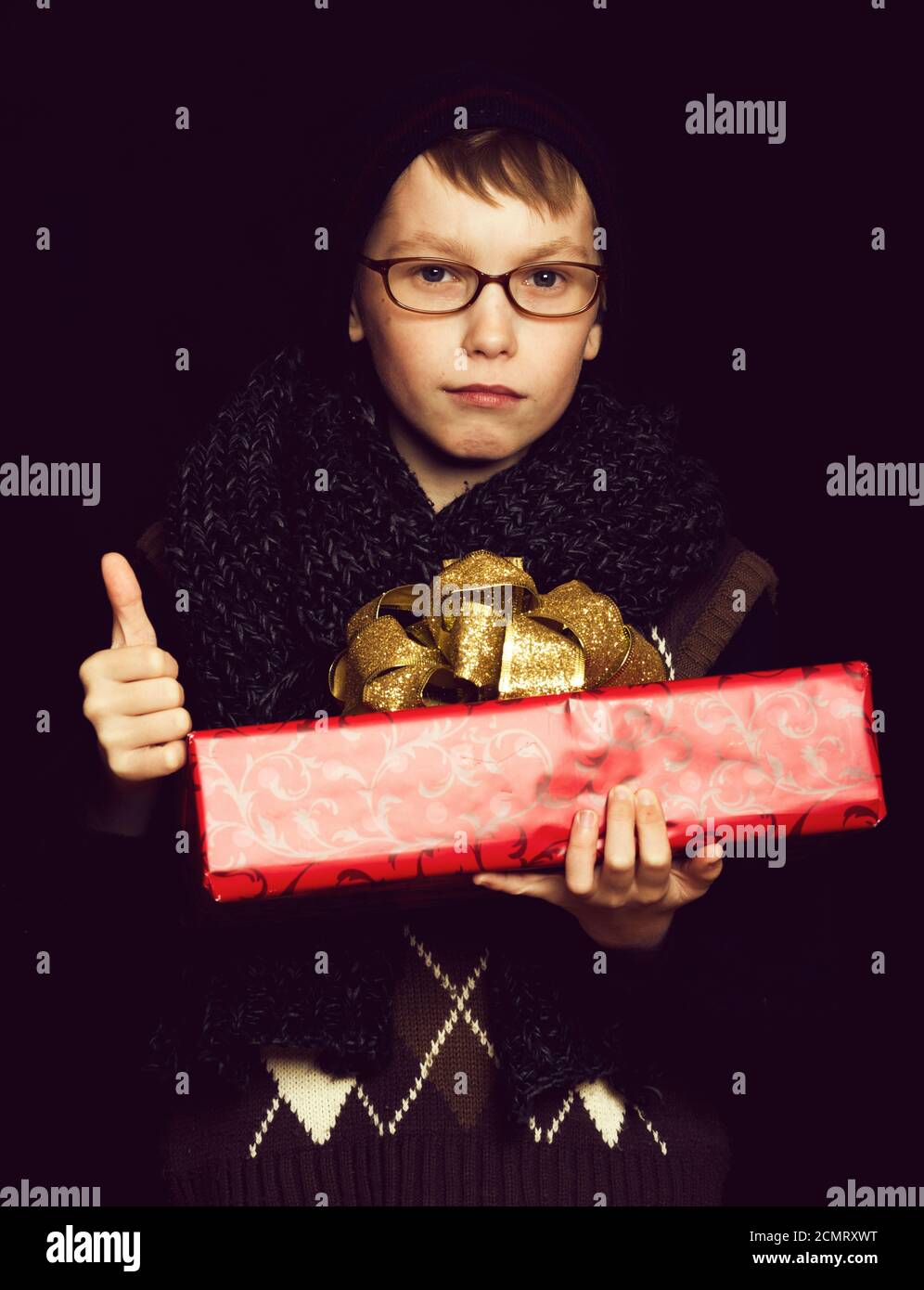 petit garçon ou mignon petit enfant nerd en lunettes, chapeau et écharpe tricotée tendance sur fond noir tient boîte cadeau rouge avec noeud doré et pouce vers le haut Banque D'Images