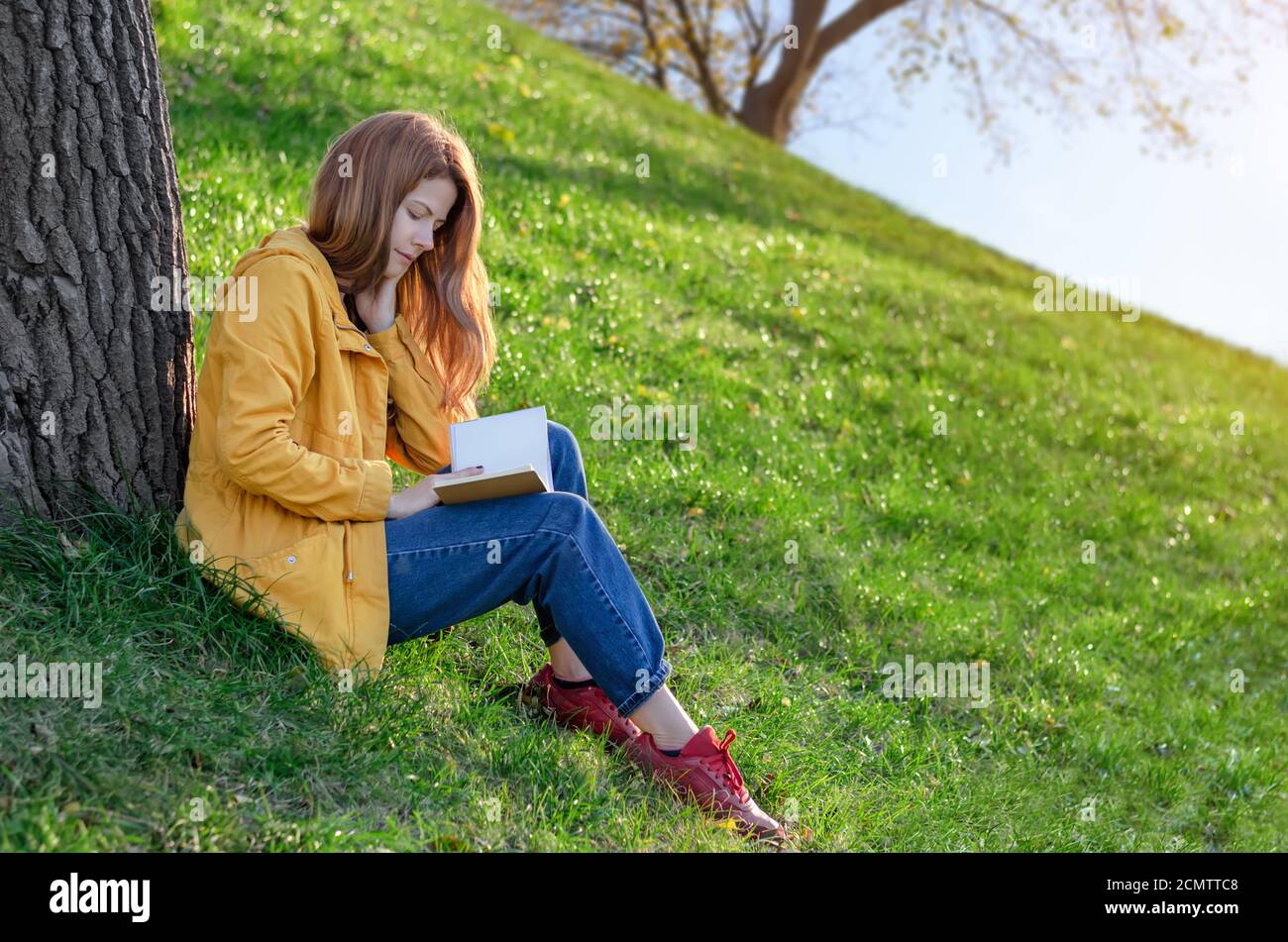 Belle jeune fille lisant un livre assis sur l'herbe verte sous l'arbre dans un parc de printemps ou d'automne Banque D'Images