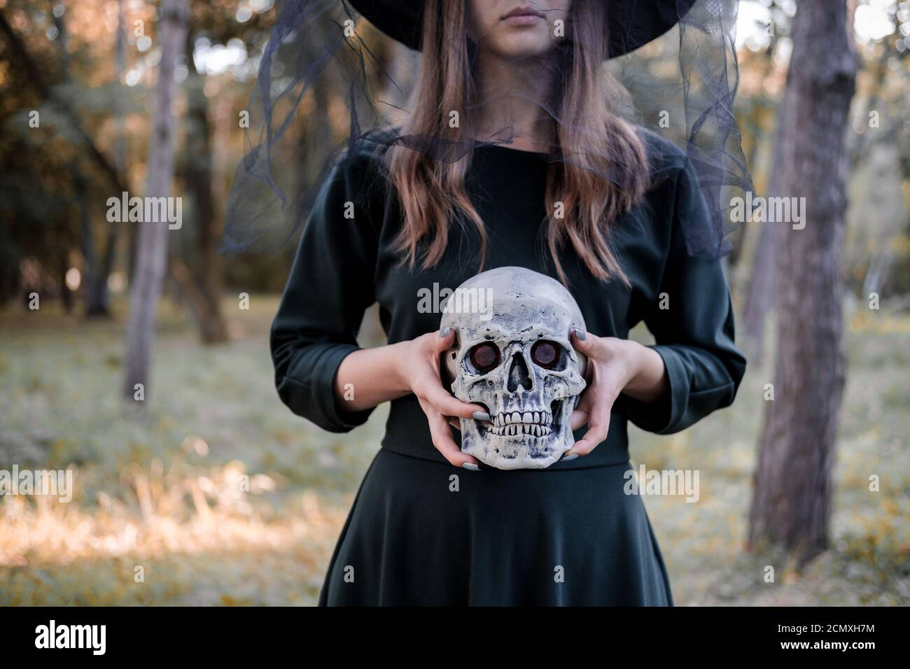 Jeune belle femme mignonne en robe sombre et chapeau de sorcière tient un crâne dans ses mains. Costume de fête d'Halloween. Forêt, parc avec arbres d'automne Banque D'Images