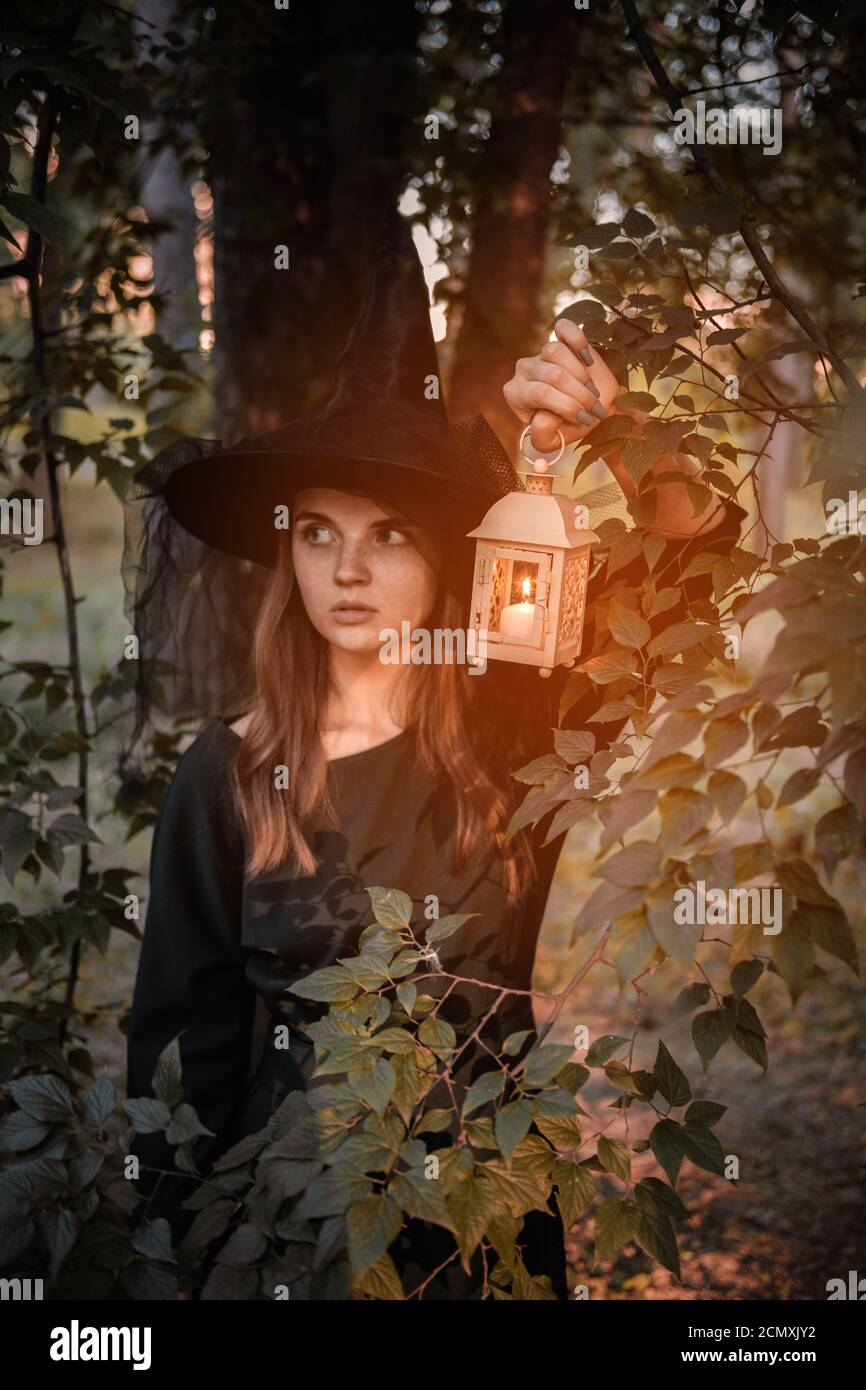 Une jeune femme en robe sombre et chapeau de sorcière tient une lanterne avec des bougies dans ses mains et illumine la forêt. Costume de fête d'Halloween. Garez-vous avec l'autom Banque D'Images