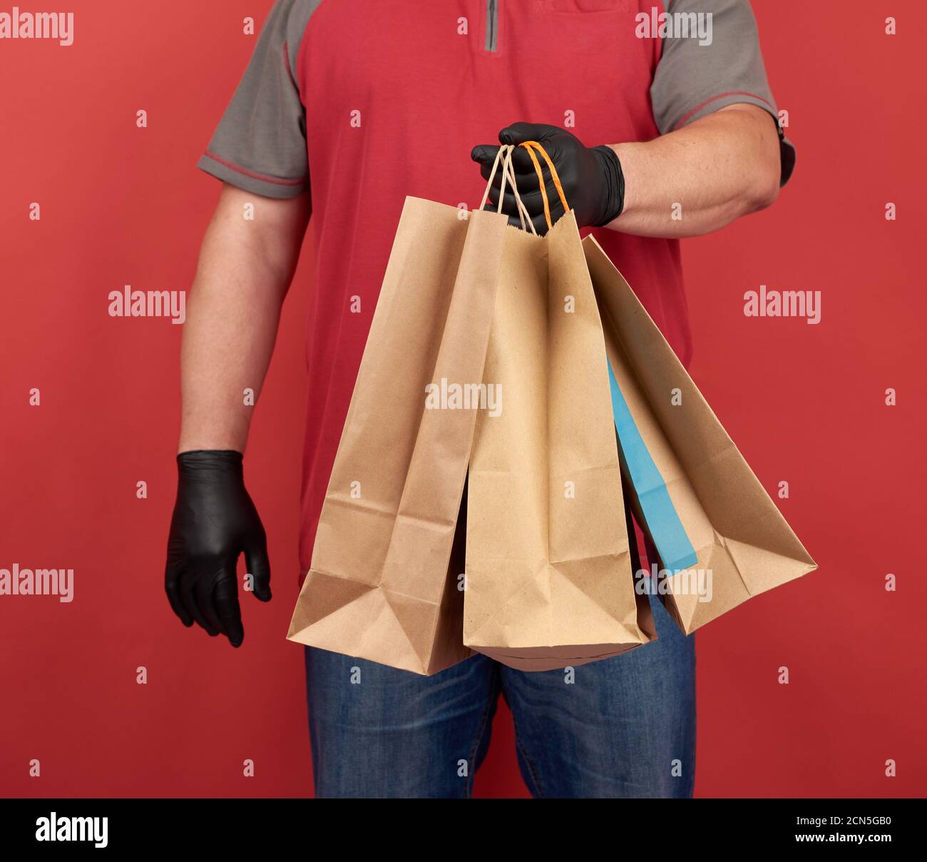 Homme dans un T-shirt rouge, portant des gants en latex noir, tient des sacs en papier pour les achats Banque D'Images