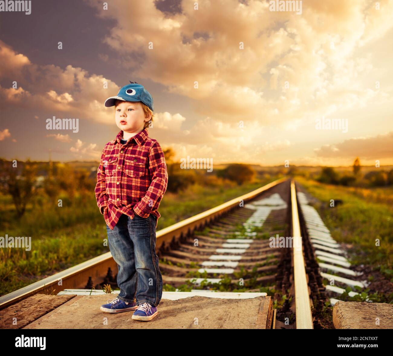 Joli petit garçon en chapeau debout dans le paysage de chemin de fer rural Banque D'Images