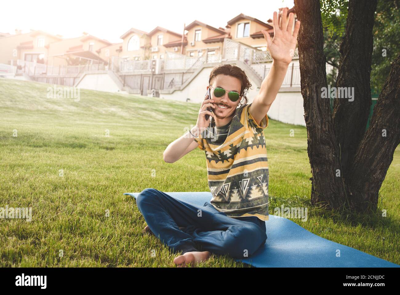 jeune homme élégant et souriant avec des lunettes de soleil est assis sur l'herbe, se reposant et parlant sur le téléphone. vagues sa main welcomi Banque D'Images