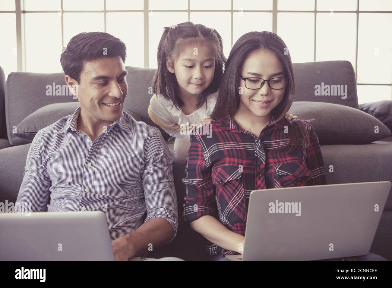 Trois membres de la famille diversifiée, le père caucasien et la mère asiatique et la petite moitié fille assis ensemble dans le salon de maison et utilisant l'ordinateur portable non Banque D'Images