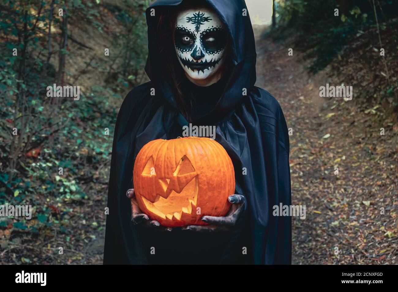 Jeune femme avec halloween peinture masque de visage portant la cagoule noire - sorcière effrayante tenant la crasse effrayante de citrouille Banque D'Images