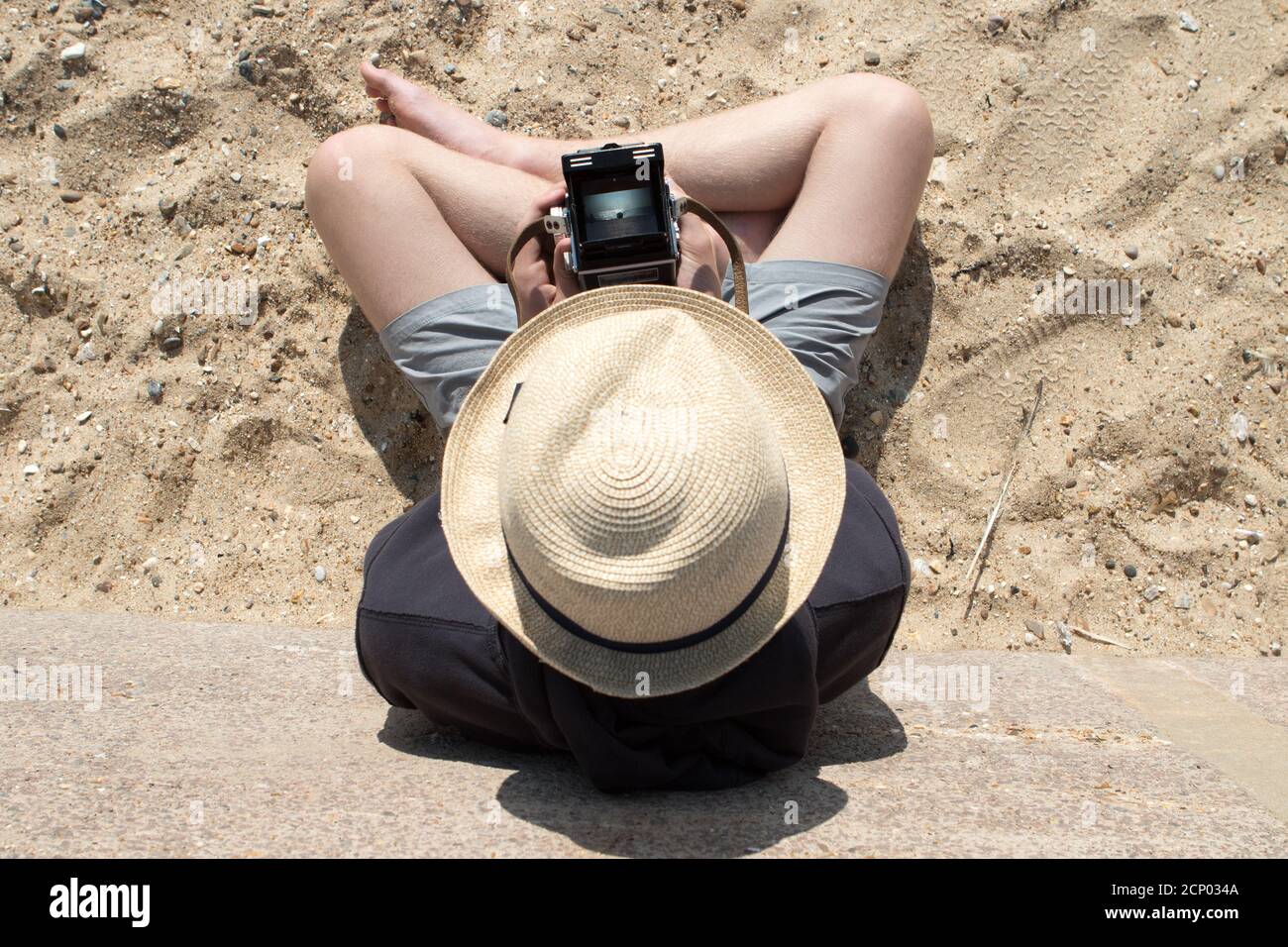 Un jeune homme adulte dans un chapeau de paille d'en haut Utilise un appareil photo reflex numérique Rolleiflex vintage pour cadrer un sujet dans une scène de plage dans le viseur Banque D'Images