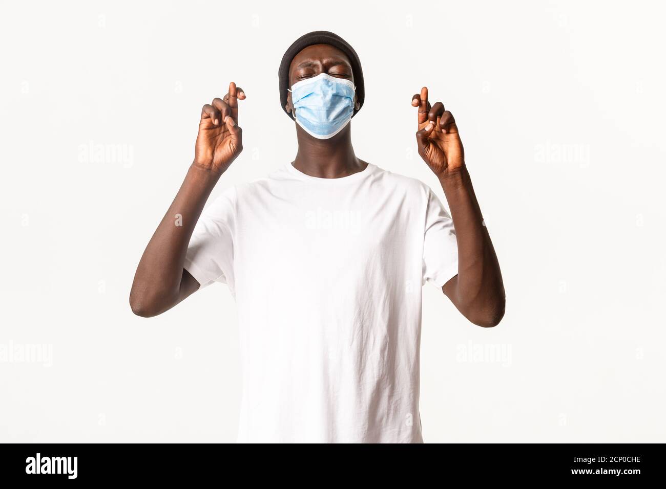 Portrait d'un homme afro-américain nerveux plein d'espoir dans un bonnet et un masque médical, croiser les doigts bonne chance et faire du voeu, debout sur fond blanc Banque D'Images