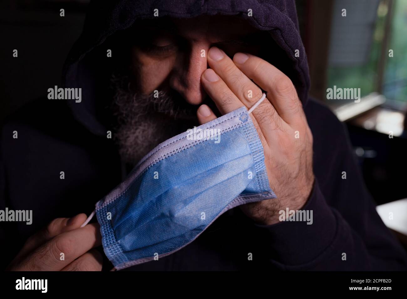 Homme barbu dans le noir à capuche tenant le masque chirurgical bleu, les yeux fermés, une main sur son visage, fatigué et déprimé regardant Banque D'Images