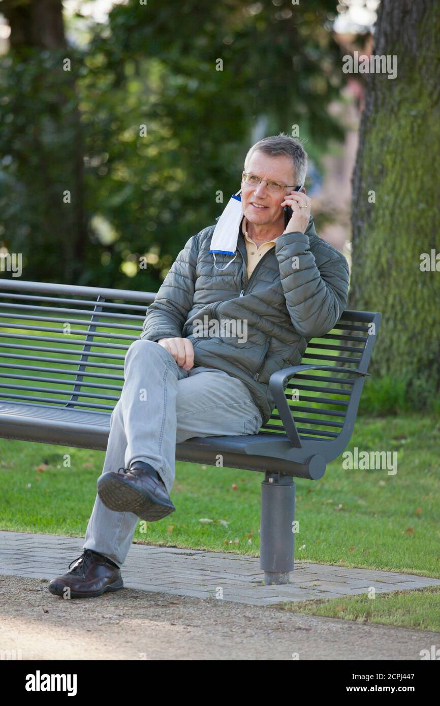 Homme mature avec masque de visage suspendu sur une oreille parlant sur un smartphone assis sur un banc de stationnement - mise au point sélective sur la tête Banque D'Images