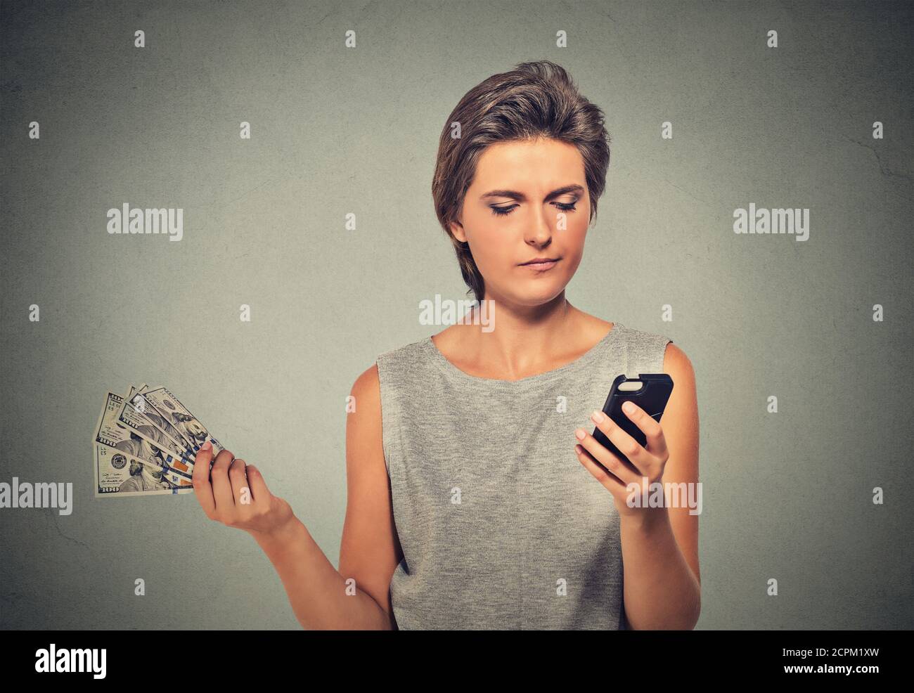 Une jeune femme malheureuse qui regarde un smartphone qui jette des billets en argent liquide sur fond de mur gris. Expression du visage contrarié. Faire des dépenses Banque D'Images