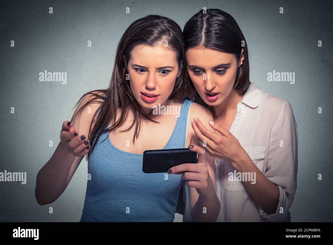 Deux jeunes filles surprises regardant un téléphone portable discutant des dernières nouvelles de potins, se sont bergées de ce qu'elles voient un fond gris isolé Banque D'Images