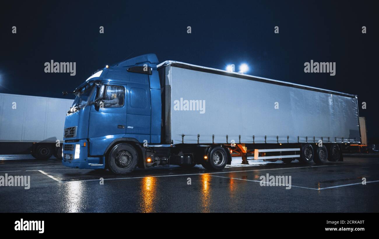 Le semi-camion bleu avec remorque de chargement se déplace dans l'espace de stationnement de nuit où les autres camions sont debout. Les conducteurs se reposant la nuit Banque D'Images