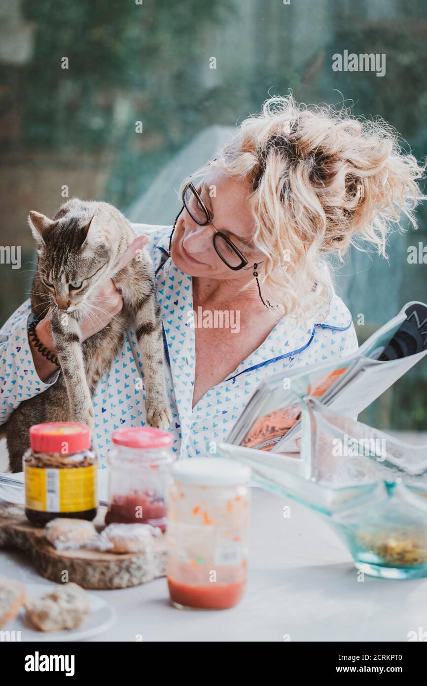 Blonde Jeune Femme Mature Avec Des Lunettes En Pyjama A La Maison A L Heure Du Petit Dejeuner Lisant Un Magazine Et Ayant Un Moment Avec Son Chat Photo Stock Alamy
