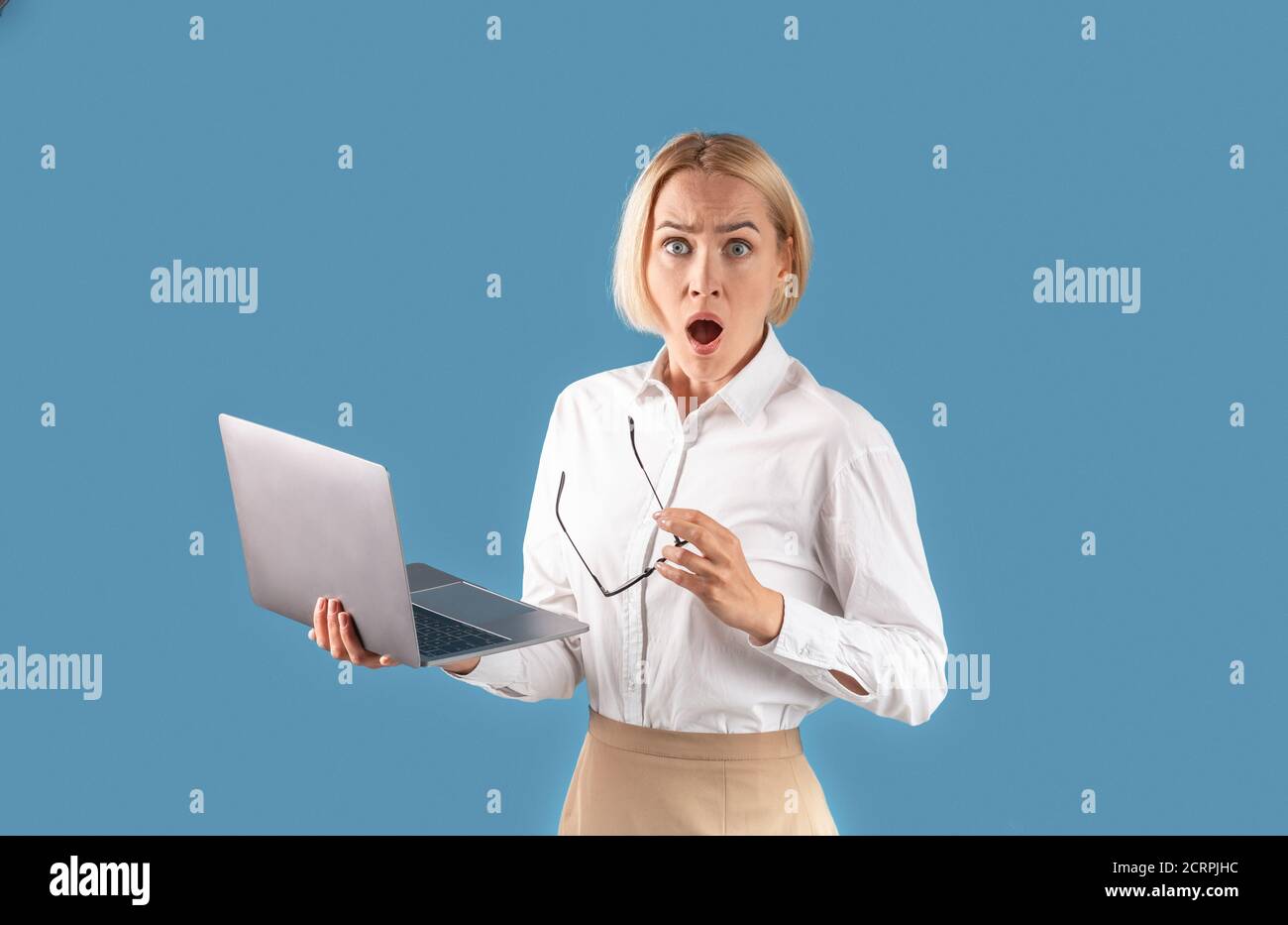 Portrait d'une femme d'affaires choquée tenant un ordinateur portable et regardant l'appareil photo sur fond de studio bleu Banque D'Images