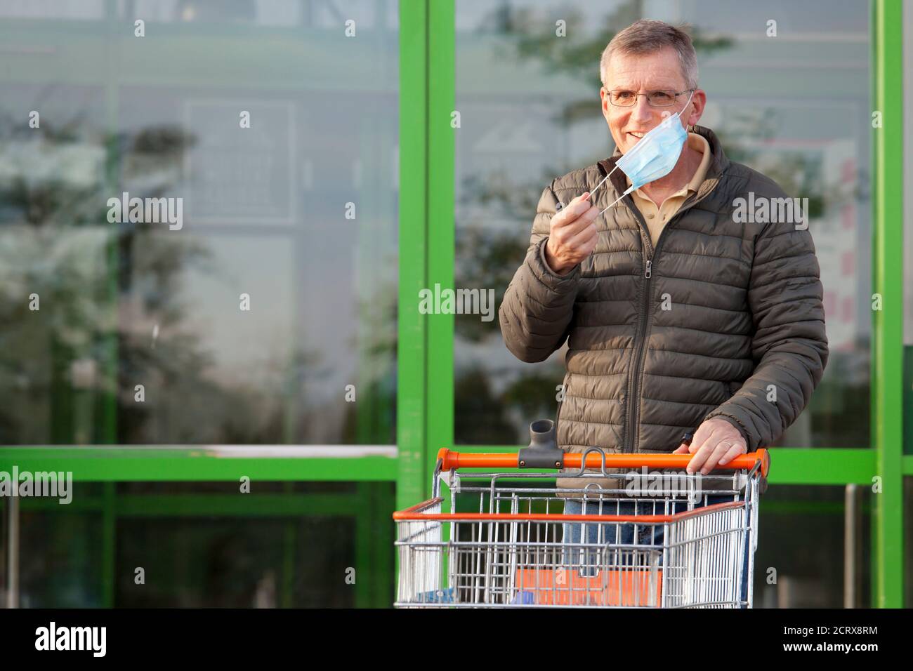 Homme mûr souriant met un masque de visage en partant un supermarché avec un panier - se concentrer sur le face Banque D'Images