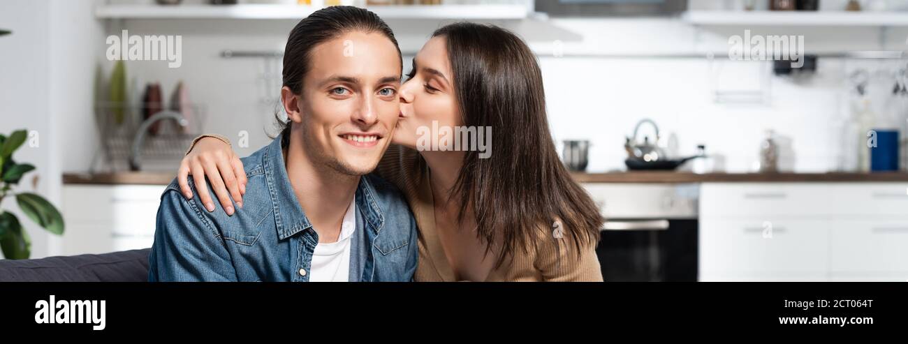 orientation panoramique de la femme brunette embrassant heureux petit ami regardant appareil photo dans la cuisine Banque D'Images