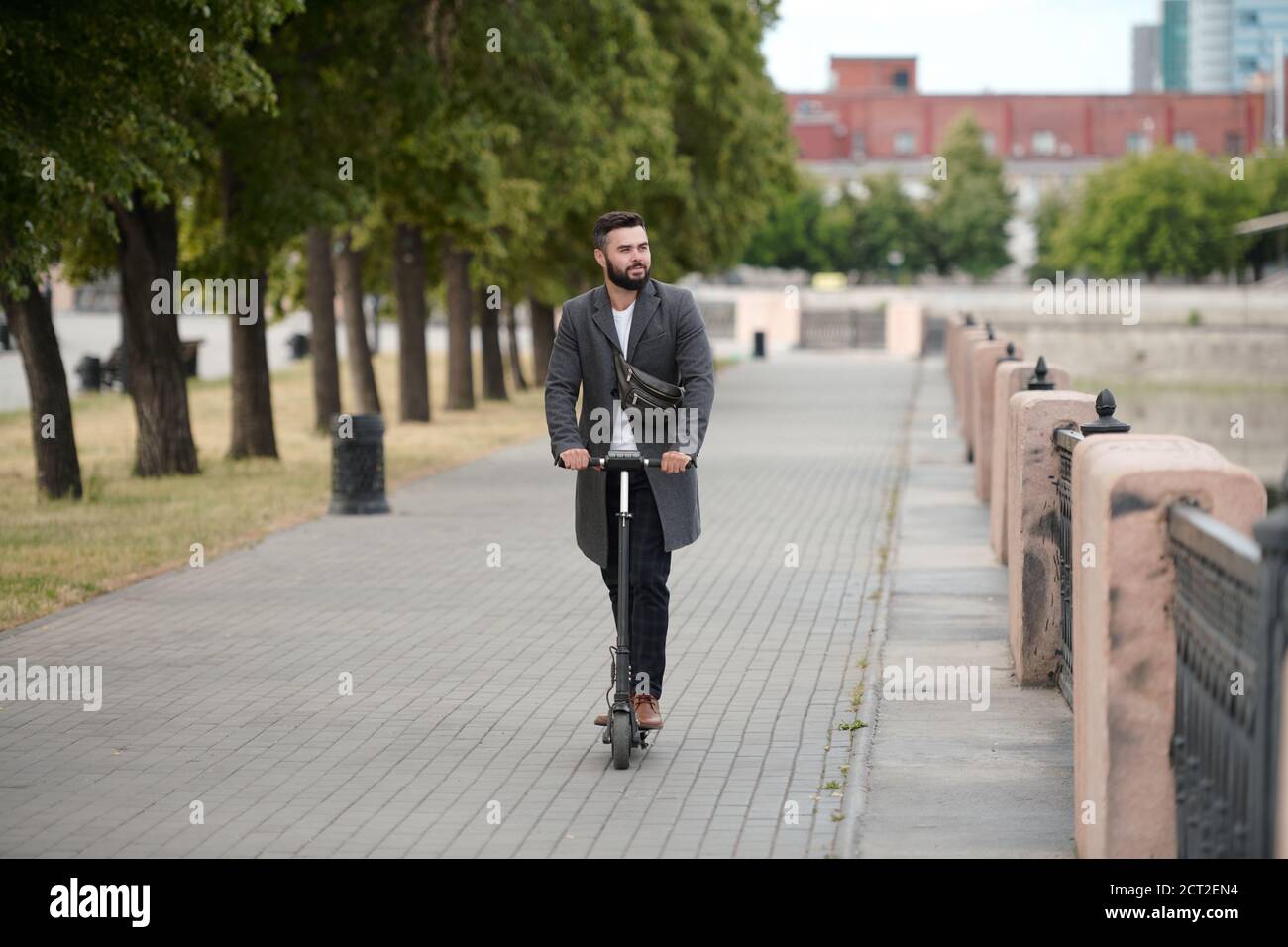 Jeune homme d'affaires contemporain barbu en manteau sur scooter électrique Banque D'Images