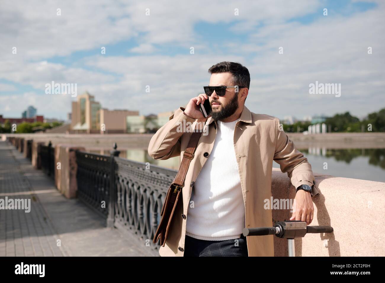 Jeune homme d'affaires barbu parlant sur smartphone tout en se tenant au bord de la rivière Banque D'Images