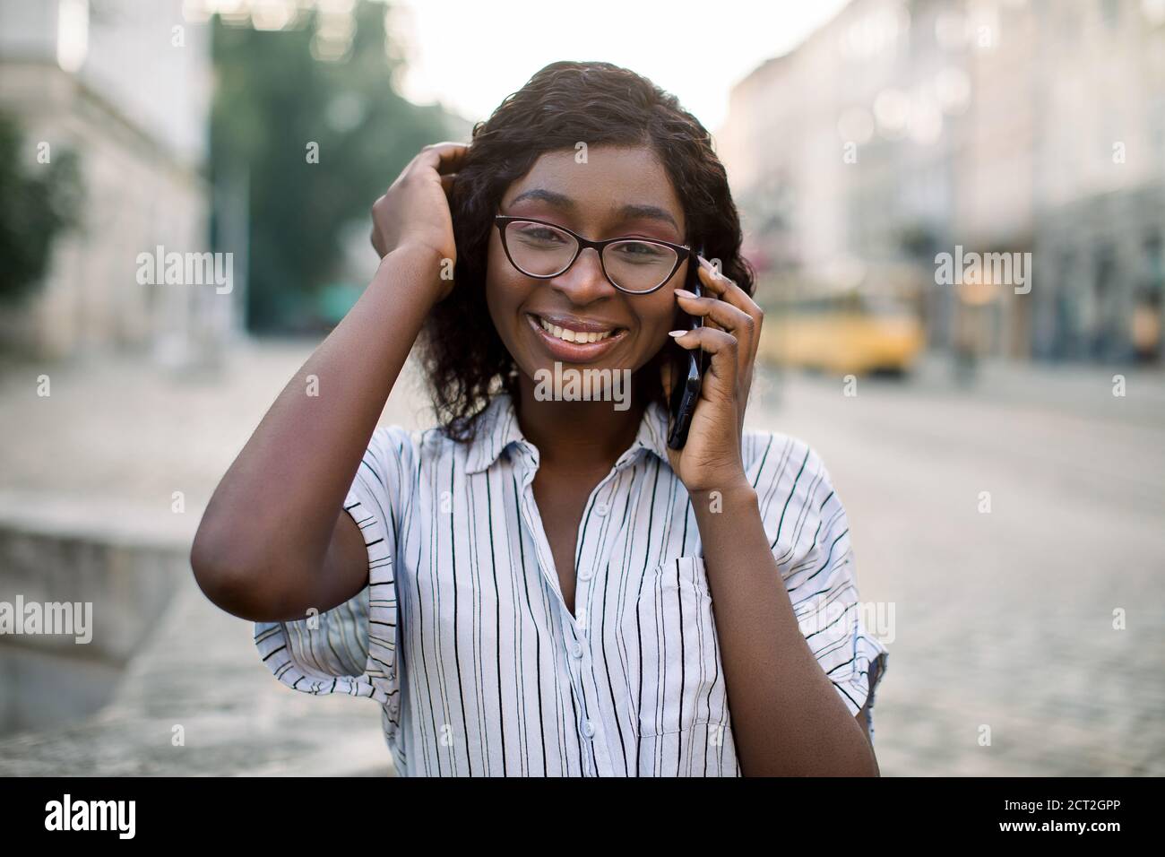 Gros plan portrait d'une femme africaine souriante en lunettes, marchant dans la vieille ville avec un téléphone portable, parlant avec son ami et souriant à l'appareil photo. Personnes Banque D'Images
