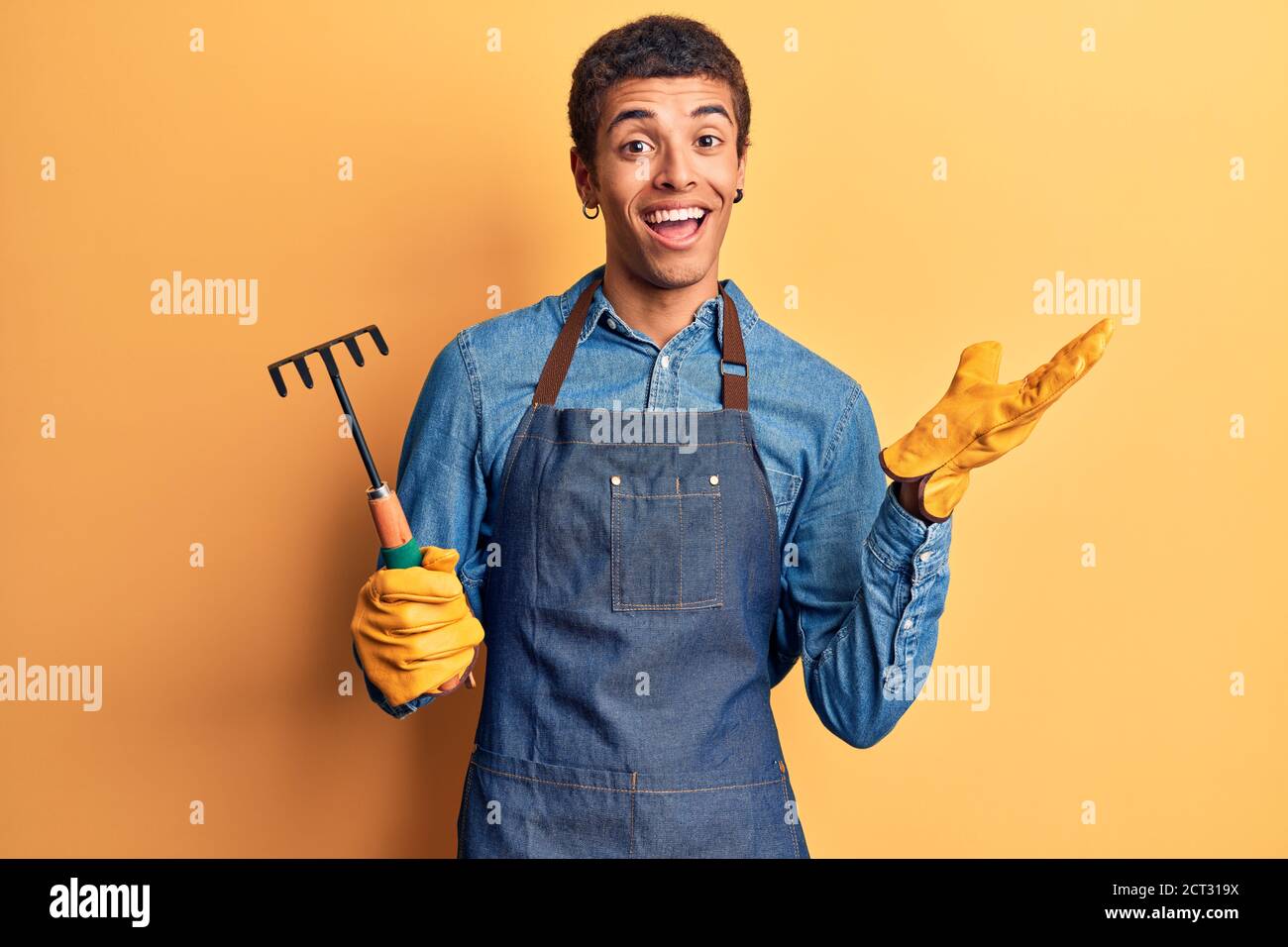 Jeune homme amérindien africain portant un tablier de jardinier et des gants tenant ratissez les succès avec un sourire heureux et une expression du gagnant avec relevé h Banque D'Images