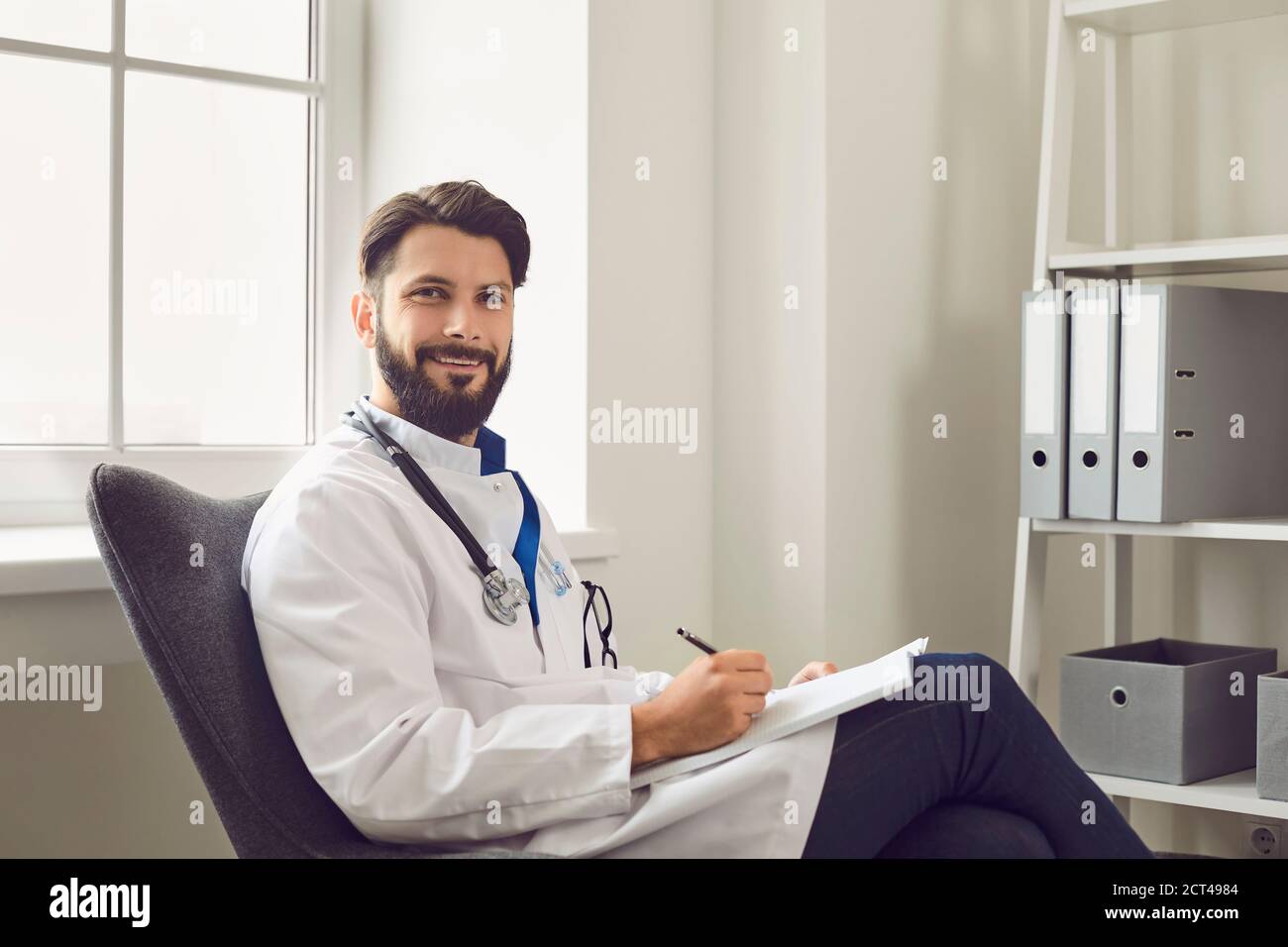 Beau jeune médecin en blouse de laboratoire assis à l'hôpital. Travailleur médical écrivant le formulaire de prescription au bureau Banque D'Images