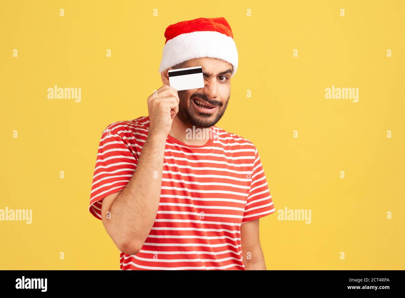 Jeune homme barbu en t-shirt rouge et chapeau de père noël se bercer et s'amuser en tenant la carte de crédit près de ses yeux, en se faisant passer. Prise de vue en studio en intérieur isola Banque D'Images
