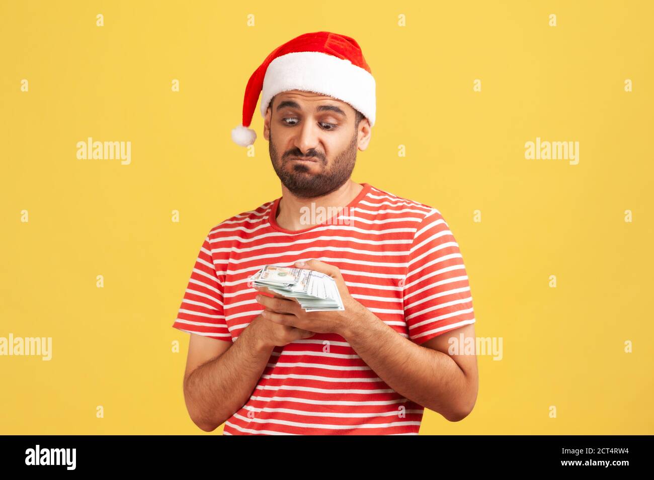 Malheureux confus barbu homme dans le chapeau du père noël tenant des dollars dans les mains, cupide pour acheter des cadeaux de vacances. Studio d'intérieur isolé sur fond jaune Banque D'Images