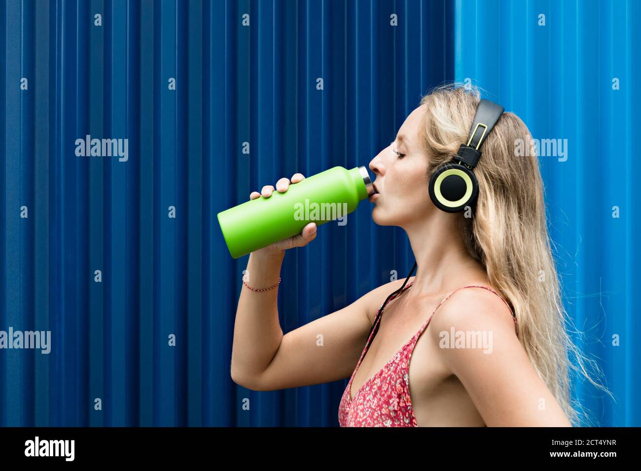 Jeune femme blonde sur fond bleu buvez de l'eau de source fraîche provenant d'un flacon vert réutilisable. Un style de vie sain en ville d'été. Banque D'Images