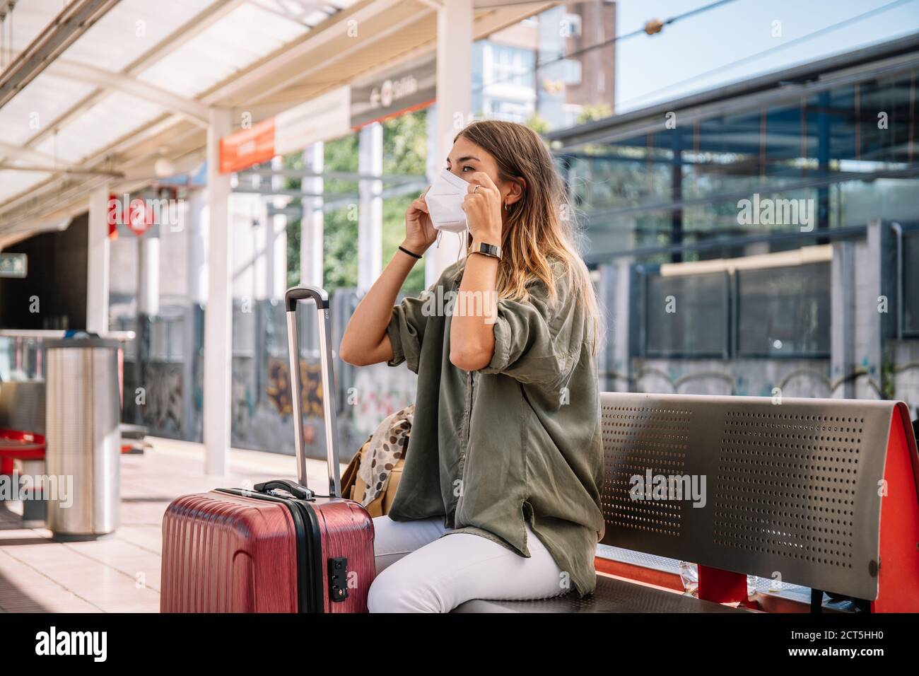 Jeune femme voyageur assis sur un banc près de la valise et de la mise sur un masque médical en attendant un train pendant l'épidémie Banque D'Images