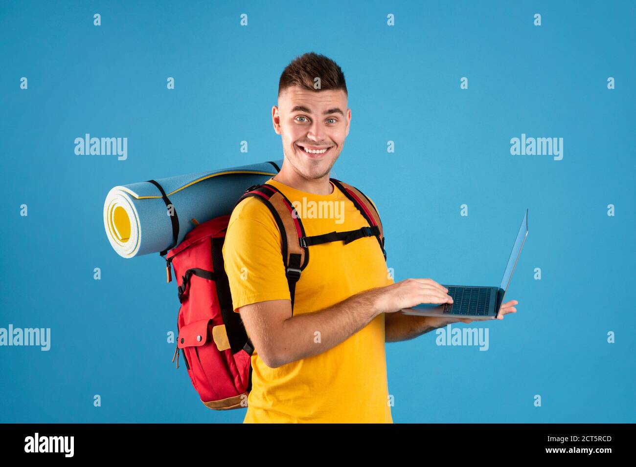 Agence de voyages en ligne. Un jeune homme heureux avec des équipements touristiques utilisant un ordinateur portable pour réserver son voyage sur Internet Banque D'Images